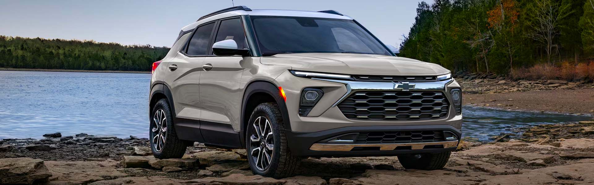 2026 Chevrolet Trailblazer compact SUV in beige with a contrasting white roof parked on a rocky lakeshore, highlighting its bold front grille, LED lighting, rugged styling, and versatile crossover design.